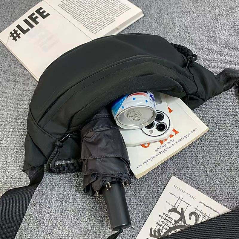 Men's black small shoulder sports crossbody chest bag with polyester fabric and nylon lining, shown with umbrella and can inside, placed on carpet with magazines.